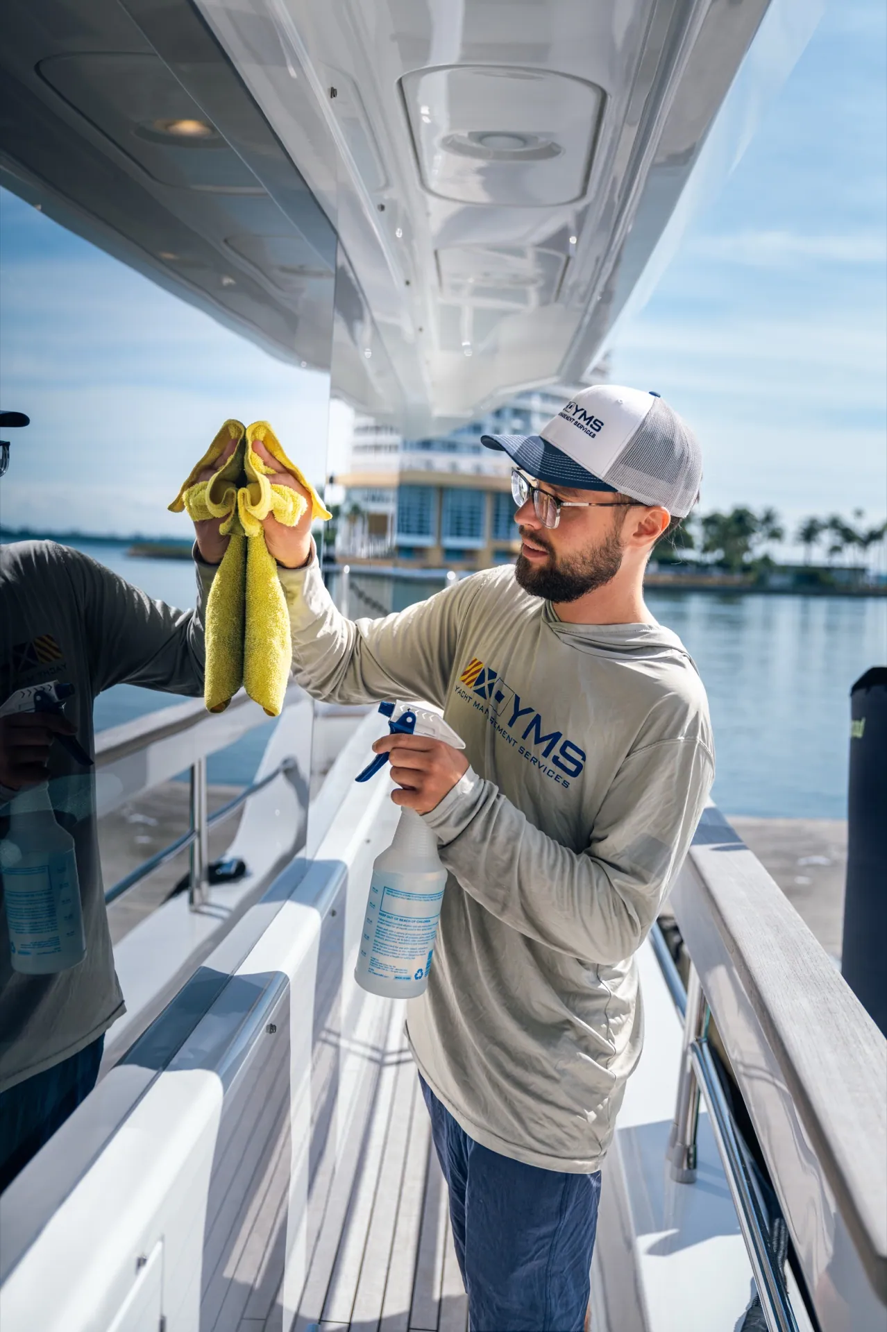A YMS crew member polishing under the hardtop of a yacht, marina and skyline behind in strong sun