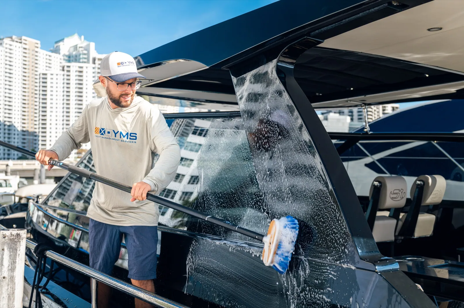 A YMS crew member sweeping a soapy brush across the windshield of a dark hulled yacht, water running down the glass, Miami towers behind