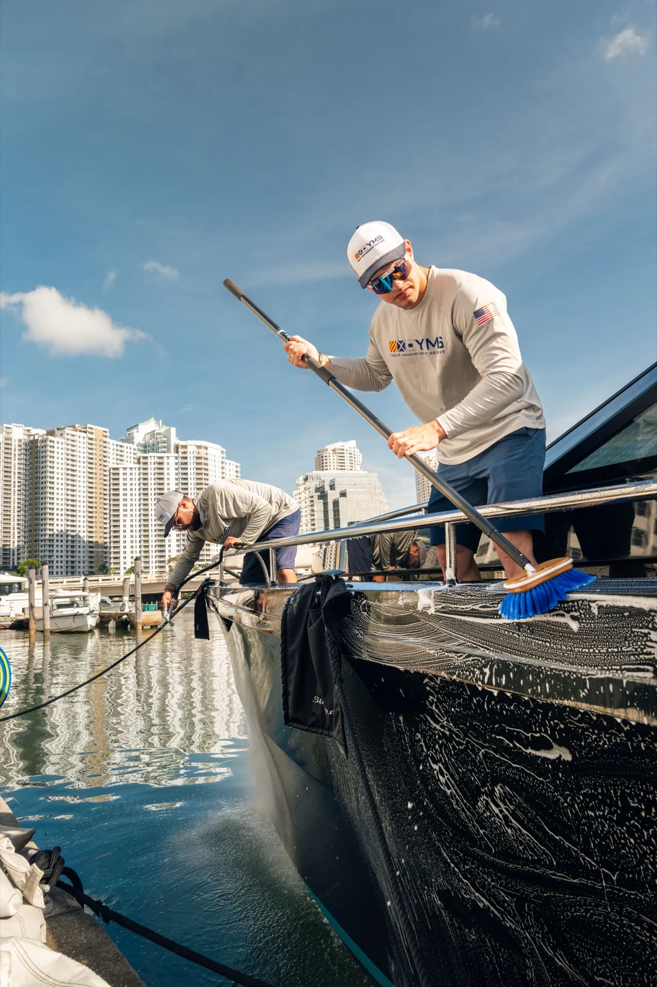 A YMS crew member reaching from the dock with a long brush down a dark hull, marina and skyline behind