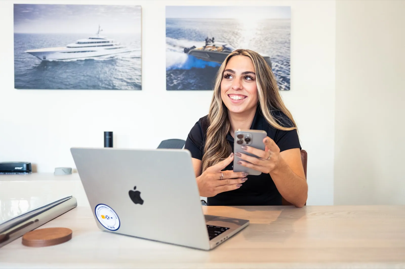 A YMS client coordinator at her desk with a phone and a laptop, yacht photography on the wall