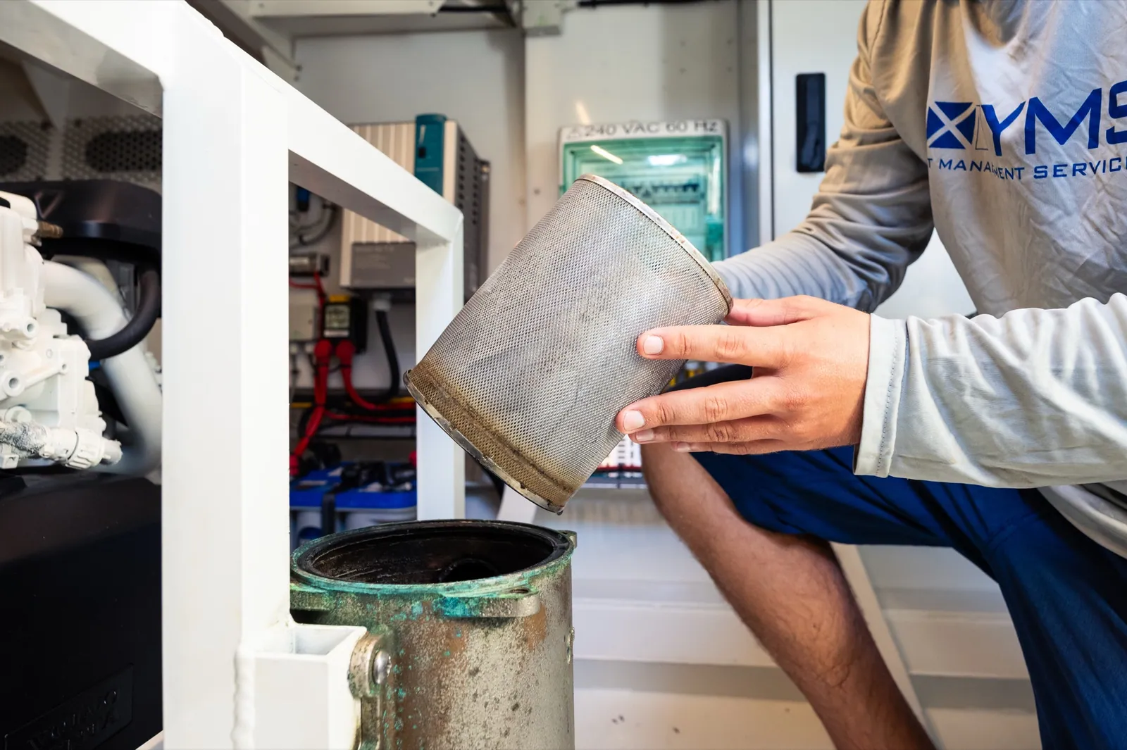 A YMS technician holding a corroded raw water strainer basket out of its housing over a bucket