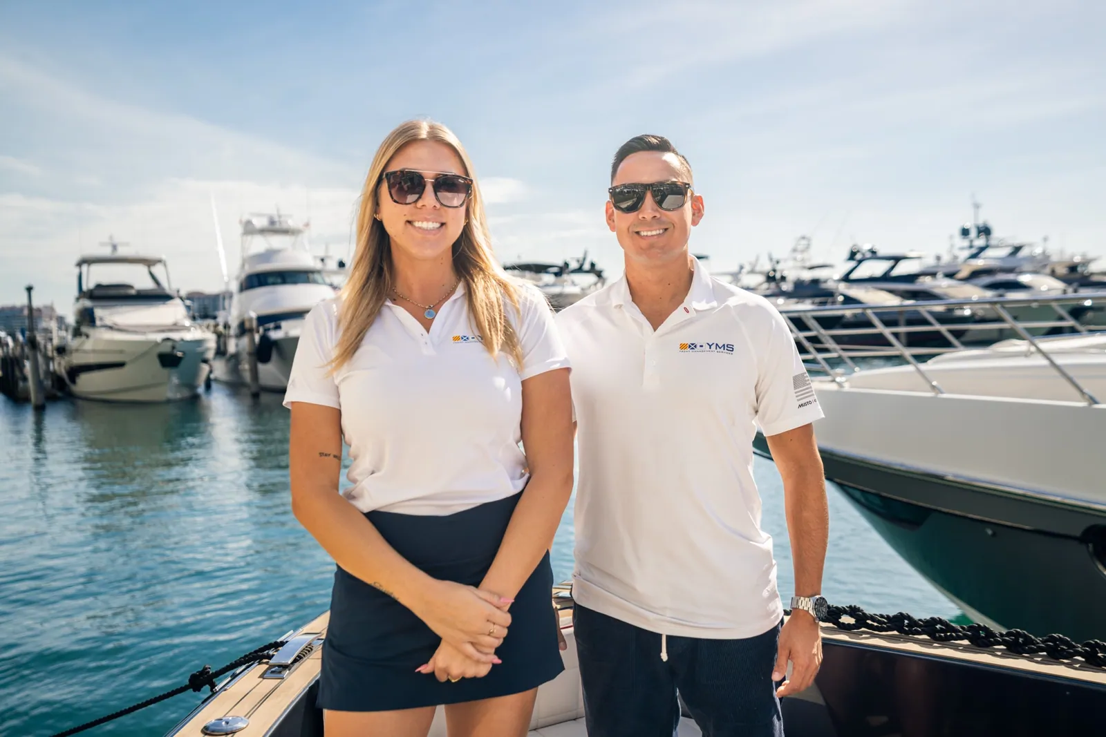 A YMS crew member and a colleague in branded polos standing at the marina, yachts behind them