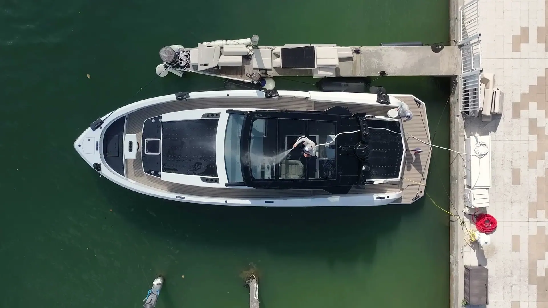 Aerial view straight down onto a white superyacht tied at a private concrete dock in flat green water, a crew member rinsing the aft deck with a hose