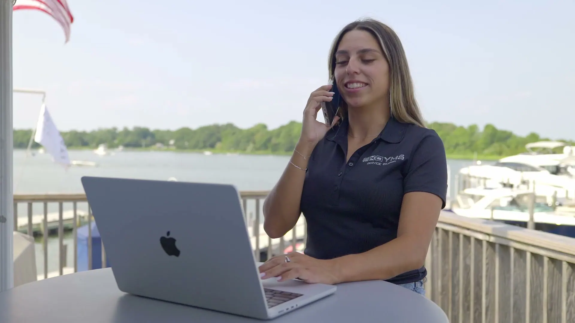 A YMS staff member on the phone at a laptop on a dock table, a marina and mangrove shoreline behind her